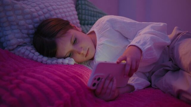 A young girl lies in bed at night under dim room lighting, looking at her smartphone screen before going to sleep.