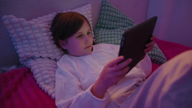 Little girl lying in bed under soft ambient lighting at night watching video content on a digital tablet before sleeping.