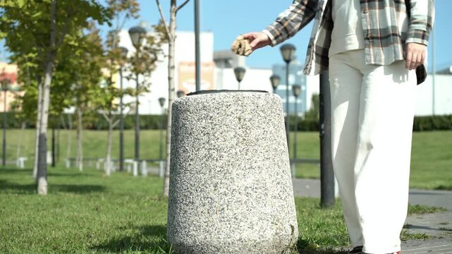 Person throwing waste into stone trash bin in sunny urban park with trees and grass. Responsible environmental action for public cleanliness and ecology.