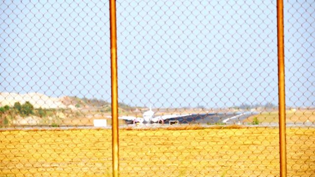 Chain-link fence dominates foreground, rusted, yellowed. Past the barrier, runway stretches under clear blue sky. White airplane sits idle, grounded, awaiting movement. Sunlight glints off the metal