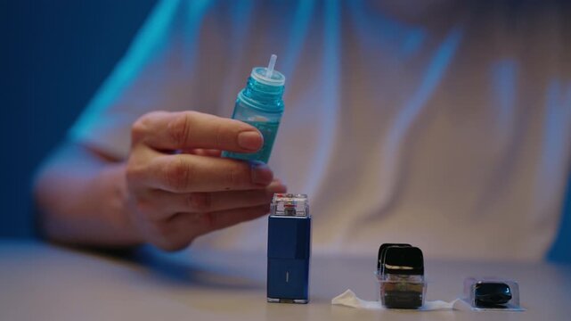 Close-up hands of unrecognizable person pouring e liquid from bottle into cartridge of pod vape system. Concept of refill process for electronic smoking device in dark room setting, slow motion.
