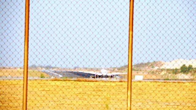 Chain fence frames distant runway. Aircraft sits idle, awaiting its next flight. Clear skies frame dry, dusty landscape. Fence rusts, weathered, standing strong. Scene hints at quiet