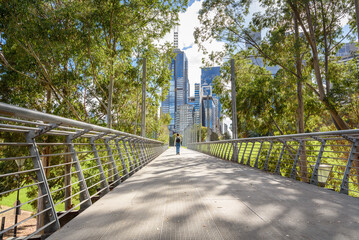 Obraz premium Woman walking on an elevated walkway lined with trees in a park bordered by modern glass office skyscrapers in a city centre on a sunny autumn day