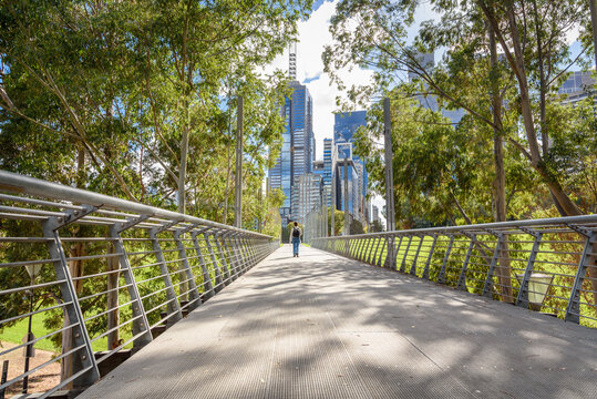 Woman walking on an elevated walkway lined with trees in a park bordered by modern glass office skyscrapers in a city centre on a sunny autumn day