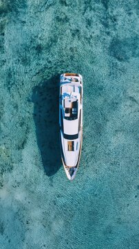 A luxury yacht floats in clear turquoise water with small sharks visible below. The aerial view highlights an elegant and exclusive yachting experience.