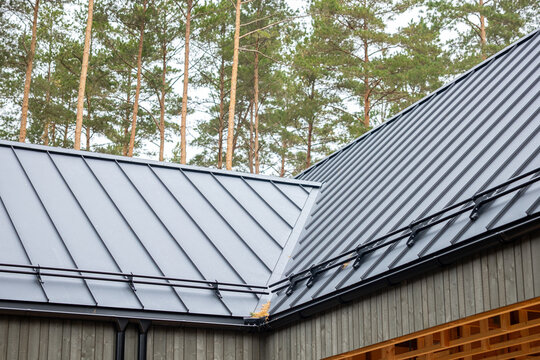 Modern metal standing seam roof of house near pine trees includes snow guards and gutters on wooden residential architecture.