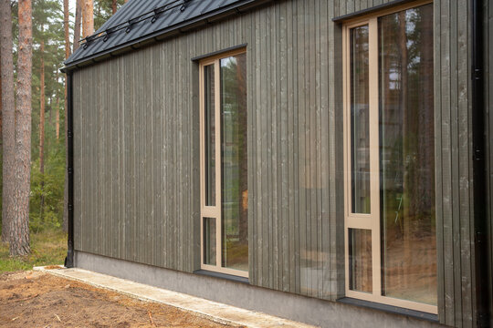 Modern wooden facade of residential house exterior displays vertical timber planks, large glass windows, and concrete foundation near pine trees.