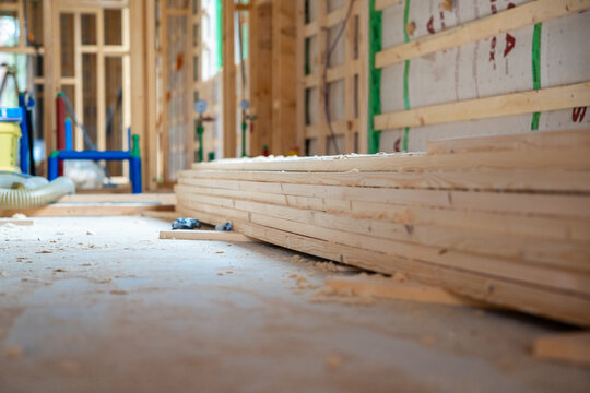 Wooden planks stacked on concrete floor inside house under construction, displaying timber framing, wall insulation, and rough utility piping at work site.