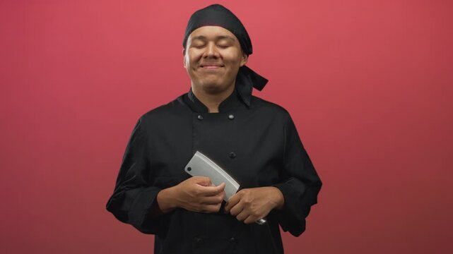 Hispanic man chef in black uniform and bandana holding a cleaver with visible hands and closed eyes in red studio; confidence craft.