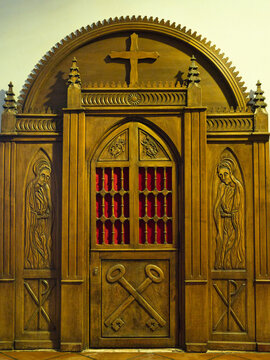 Ornate Wooden Church Confessional Door - Intricately carved wooden confessional door featuring religious symbols like a cross, saints, Chi-Rho, and crossed keys, set against a red f