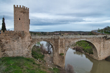 Obraz premium Historic Bridge and Tower Over Tagus Riv - Panoramic view of the iconic Puente de San Martín, a medieval arch bridge, and its fortified tower spanning the Tagus River in Toledo, Spain