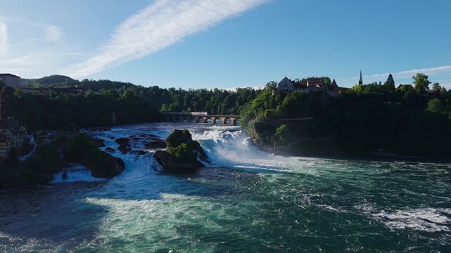 Forward and upward drone flight over the Rhine Falls in Schaffhausen, featuring river rapids, a historic castle, and a train crossing the bridge.