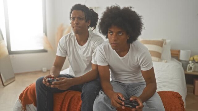 Two men sit on a bed holding gaming controllers, hands gripping joysticks, leaning forward and concentrating in a building; friendly competition.