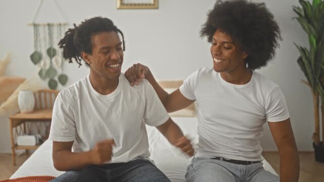 Two men on bed laugh, one with hand on shoulder while they exchange a playful fist bump in a building; joyful camaraderie.