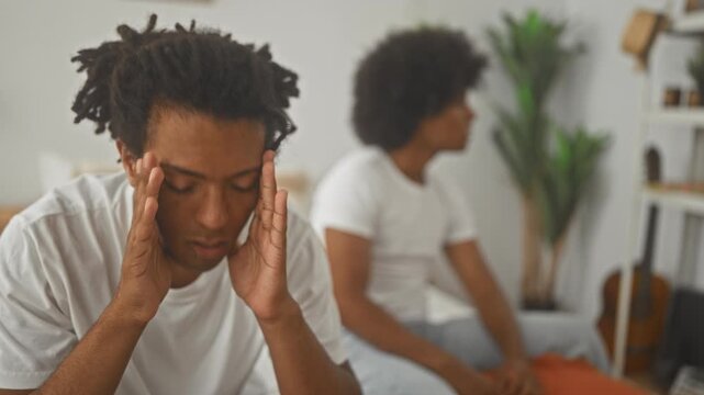 Man face downcast seated on bed in bedroom while friend gestures with hand and speaks; relationship tension.