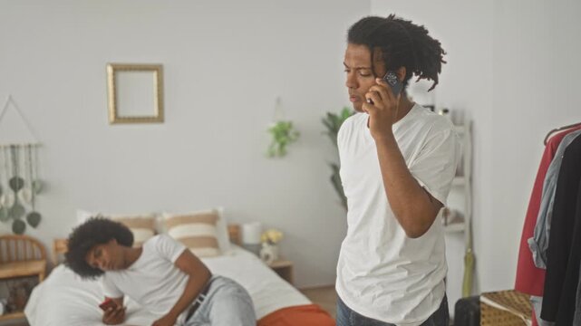 Man stands holding smartphone to ear in a bedroom inside a building, near bed while friend reclines and scrolls on phone; concern conversation.
