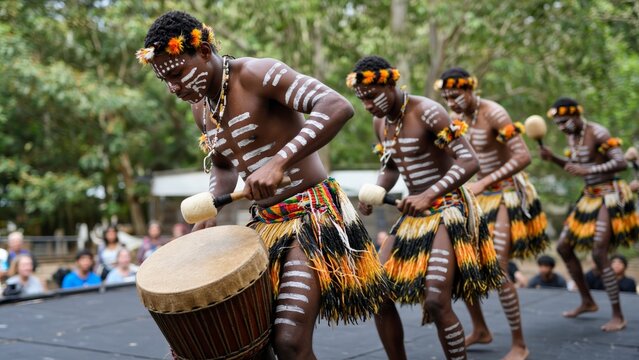 Traditional performers beat drums in outdoor cultural performance. Body painted men move in coordinated rhythm, wearing colorful skirts and headbands, while audience watches from behind stage.
