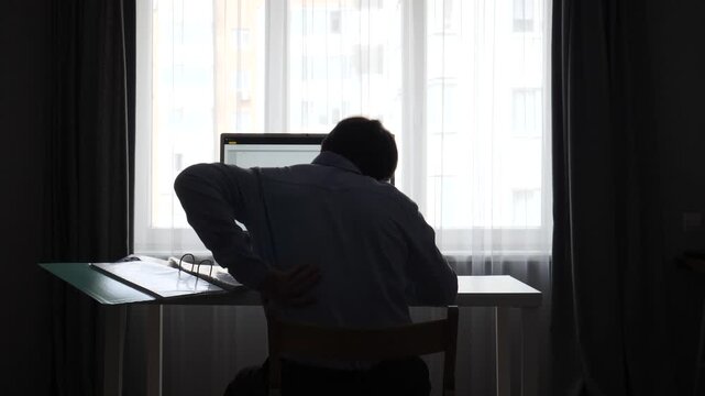Man feeling discomfort and rubbing his lower back while sitting at a home office desk, experiencing pain from prolonged sedentary computer work in silhouette