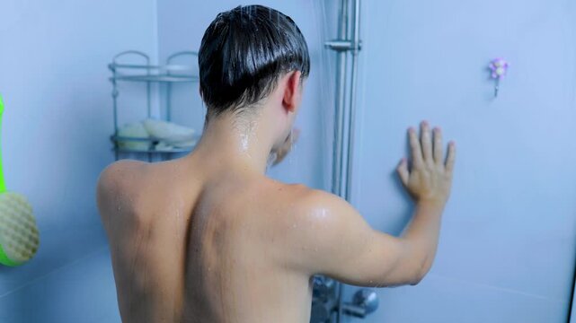Tired teenage guy leaning on the home bathroom wall under a refreshing shower.