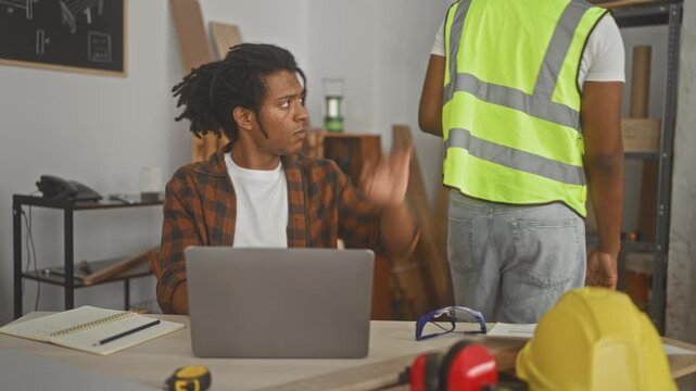 Two carpenter partners consult a laptop as one leans over pointing at the screen in a workshop building; focused teamwork.