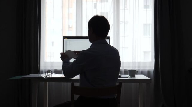 Man in silhouette experiencing frustration and stress while working at a desk with a computer, tearing up documents in his home office during remote work