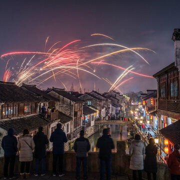 People watching a vibrant fireworks display over an ancient Chinese water town canal at night