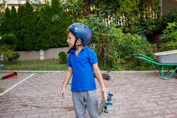 Fototapeta premium Young boy wearing helmet holding skateboard in backyard, brick paved surface, green trees and bushes background, wheelbarrow nearby, residential setting, high quality photo