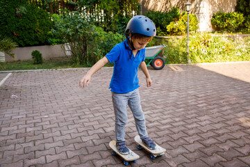 Fototapeta premium Young boy wearing safety helmet skateboarding in backyard, brick paved surface, garden with potted plants and wheelbarrow, residential buildings in background, active childhood outdoor recreation
