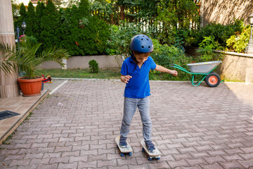 Fototapeta premium Young boy wearing safety helmet skateboarding in backyard, brick paved surface, garden with potted plants and wheelbarrow, residential buildings in background, active childhood outdoor recreation