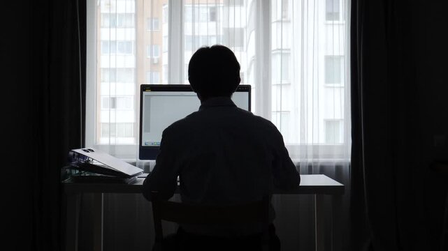 Man sitting at a desk in a dark room, working on a computer backlit by a window with sheer curtains and an urban building view, representing remote work and professional productivity