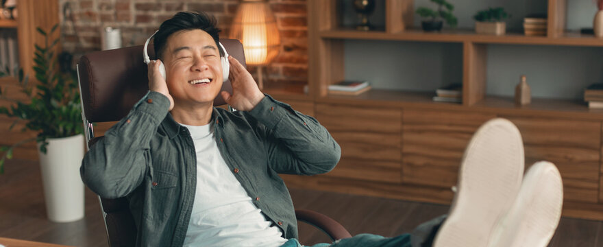 Happy Asian Male In His 20s Listening To Music With Headphones While Relaxing In A Cozy Home Office, Feet Up In Chair, Conveying Comfort And Enjoyment In A Modern Setting. Home Lifestyle Concept.
