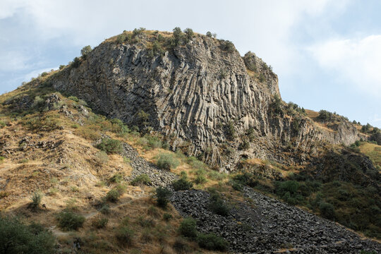 Layered basalt rock formation in Armenia, featuring greenery and rocky ground, with a cloudy sky providing a dramatic backdrop to the landscape