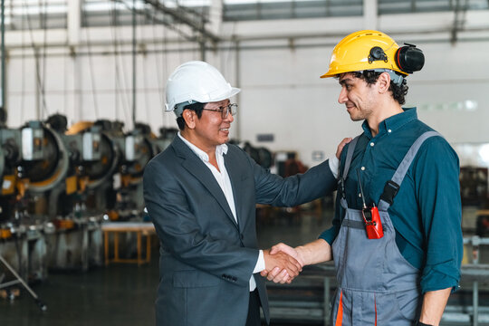 Asian manager and male industrial worker shaking hands inside factory, expressing trust and professional cooperation during workplace discussion
