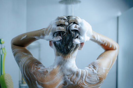 Rear view of a young guy washing his back with cleansing foam indoors.