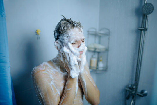 Adolescent guy cleanses his face with thick foam in the home shower.