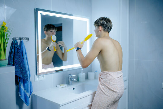 Happy adolescent guy singing holding yellow bath brush facing bathroom mirror.