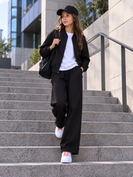 Stylish teenage girl in black sportswear and cap walking down urban stairs outdoors. Concept of pal, streetwear, urban fashion, baseball cap, casual outfit, teenager, full length.