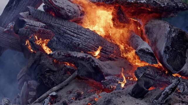 Fire with charring and smoke. Destructive wood combustion beneath heavy smoke. Blackened logs during combustion with orange flames. Combustion damage to wood and branches.