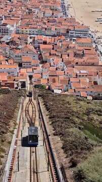 Two Nazar&eacute; funicular cars cross on the hillside, with one ascending while the other simultaneously travels in the opposite, descending direction toward the town and beach in Portugal.