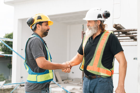 Construction worker handshake at building site shows teamwork, safety equipment, and professional cooperation during outdoor renovation work with unfinished structure and tools nearby