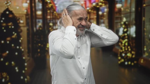 Man with gray hair and beard covers ears in decorated shopping building corridor holding hands to head in pain; holiday discomfort.