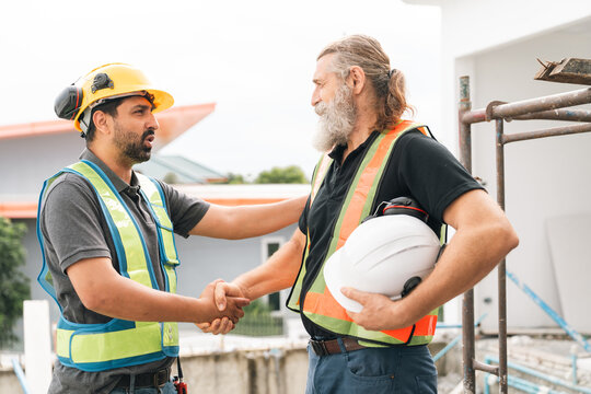 Construction worker and supervisor shaking hands beside building project, wearing safety helmet and reflective vest, with white hard hat under arm and friendly teamwork expression