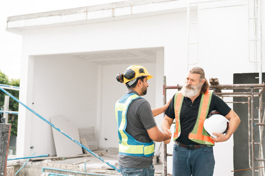 Construction worker safety vest hard hat handshake building site Construction worker wearing safety vest and hard hat shakes hands with bearded supervisor building site, holding helmet during