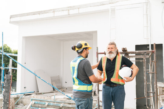 Construction worker and site supervisor discuss renovation progress beside modern building, wearing safety vest and helmet while holding hard hat outdoors in daylight