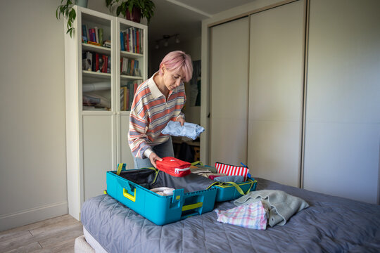 Focused woman preparing for holiday trip and attentively packing suitcase on bed in bedroom, concentrated female traveler organizing personal things in luggage, placing necessary stuff and accessories