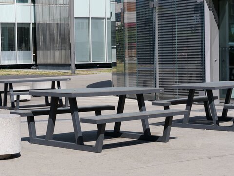 Outdoor corporate campus amenities: Modern dark gray picnic tables and benches on concrete ground near contemporary glass office buildings in Germany.