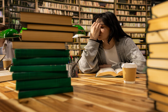 Tired young woman struggles with an open book during exam preparation in a university library, showing stress, fatigue and burnout during coursework.