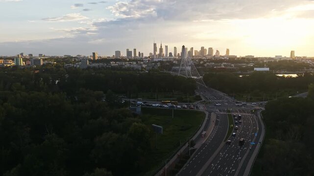 Forward drone flight toward Warsaw city center at golden hour, revealing downtown skyline with modern skyscrapers, busy multilane road, moving traffic, green parkland and cable-stayed bridge
