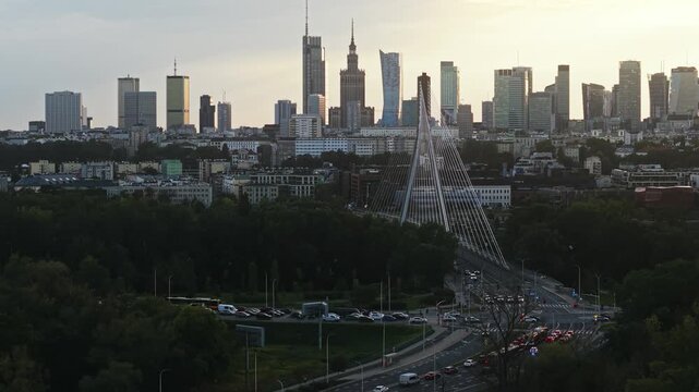 Forward drone flight toward Warsaw city center at golden hour, revealing downtown skyline with modern skyscrapers, busy multilane road, moving traffic, green parkland and cable-stayed bridge