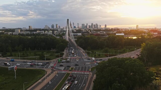 Smooth aerial panorama from right to left over Warsaw at golden hour, revealing Swietokrzyski Bridge, busy urban intersection, moving traffic, green parkland and modern downtown skyline